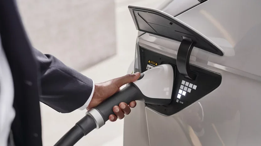 A hand in a dark suit is plugging a white charging nozzle into the port of a light-gray electric car.